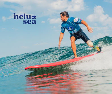 Inclusea adaptive surfing image of a man wearing a shortie wetsuit and blue rash vest with a prosthetic leg surfing an unbroken wave on a red surfboard.