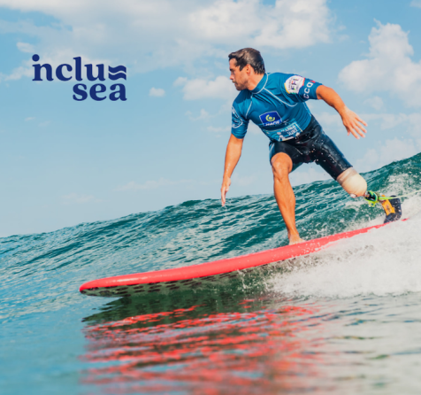 Inclusea adaptive surfing image of a man wearing a shortie wetsuit and blue rash vest with a prosthetic leg surfing an unbroken wave on a red surfboard.